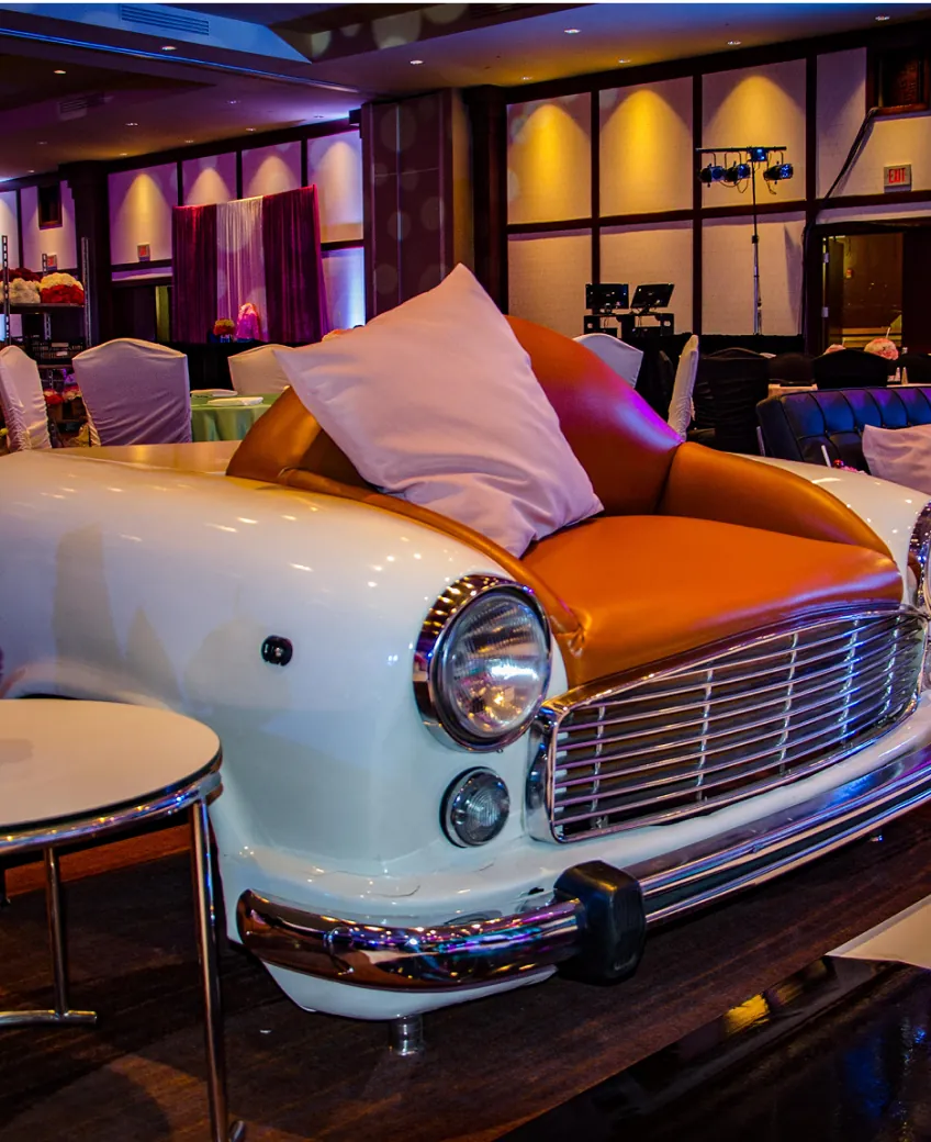 A vintage white car turned into a lounge seat, with an orange leather cushion, at a themed party