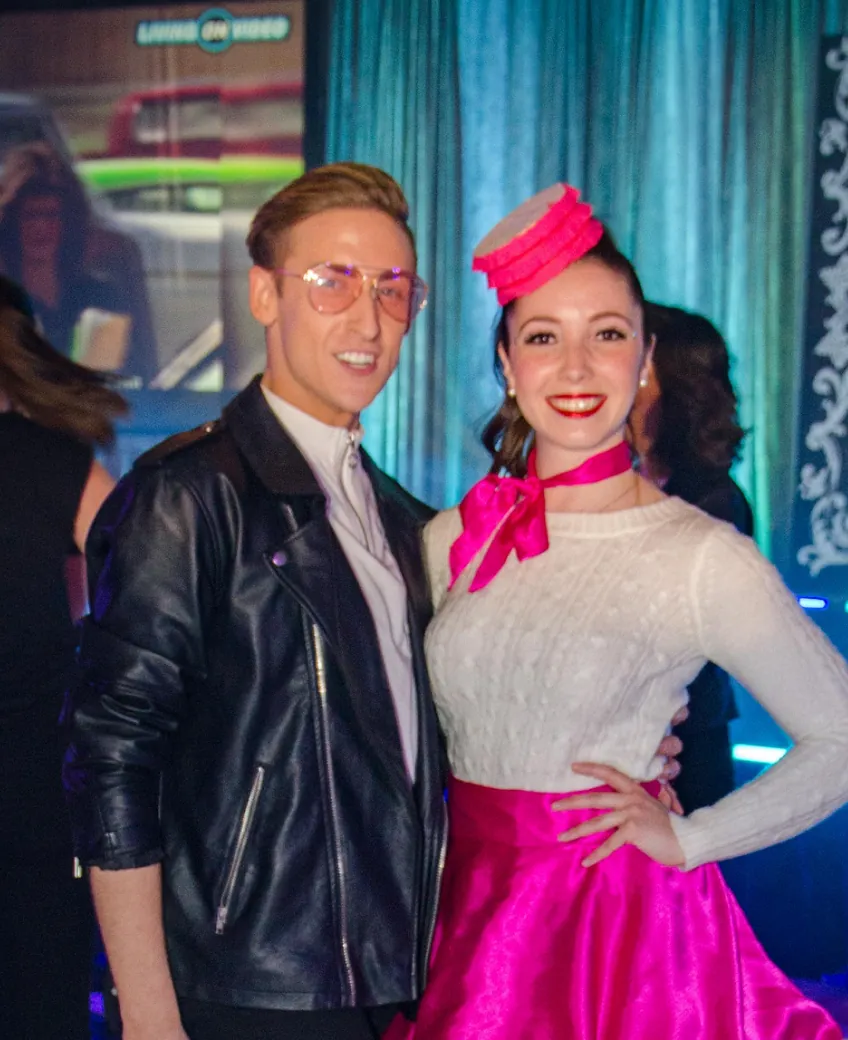 Two guests in 1950s costume, leather jacket and pink poodle skirt, in front of a teal curtain